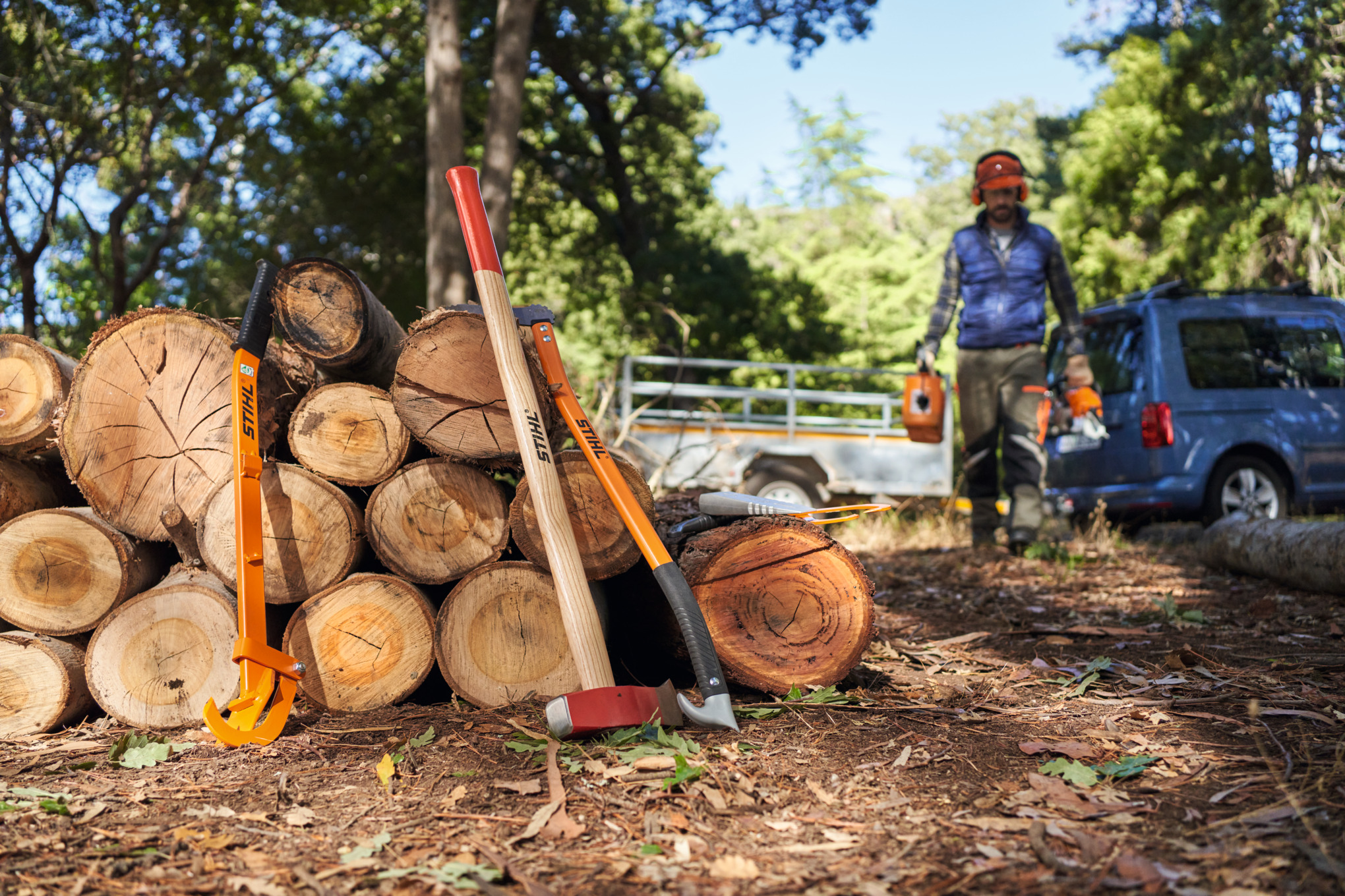 Muškarac u zaštitnoj opremi i sa uređajima u rukama približava se gomili drva za ogrev na koju su naslonjeni STIHL sekira i drugi uređaji za cepanje drva