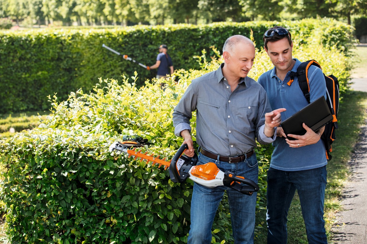 Dr Nikolas Stihl drži u ruci STIHL trimer za šišanje žive ograde i razgovara sa osobom koja stoji pored njega i drži laptop.