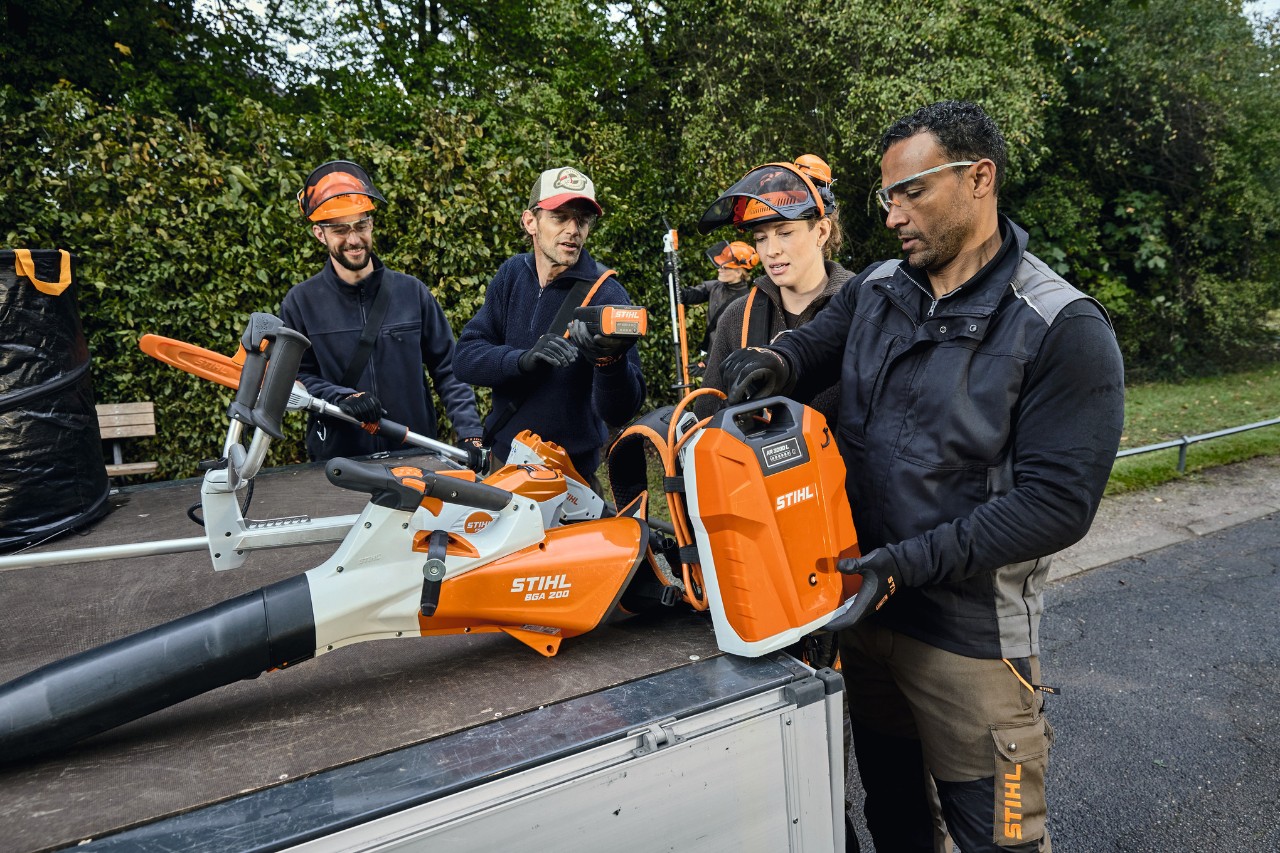 Tri muškarca i jedna žena u STIHL zaštitnoj opremi stoje na tovarnoj površini vozila sa STIHL uređajima kao što su, na primer, baterijski duvač lišća STIHL BGA 200 i leđna baterija STIHL AR 2000 L 