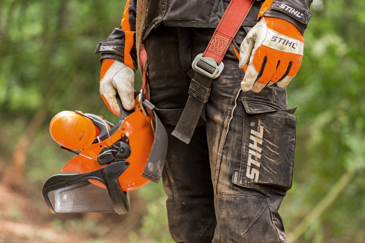 Osoba u STIHL pantalonama sa zaštitom od prosecanja i STIHL rukavicama drži u ruci STIHL zaštitni šlem 
