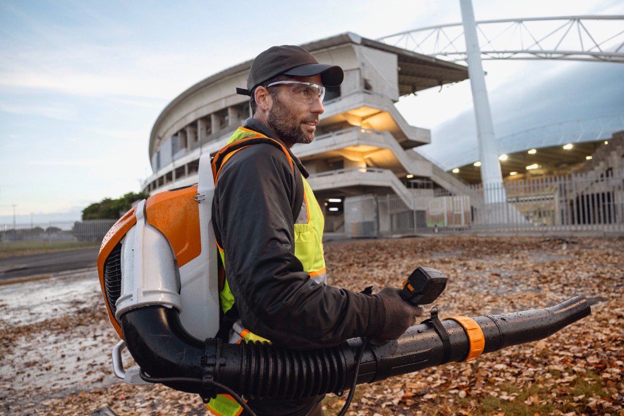 Kasno uveče osoba čisti lišće i ostatke sa stadiona koristeći bežični puhač lišća ALLPRO STIHL BRA 600.