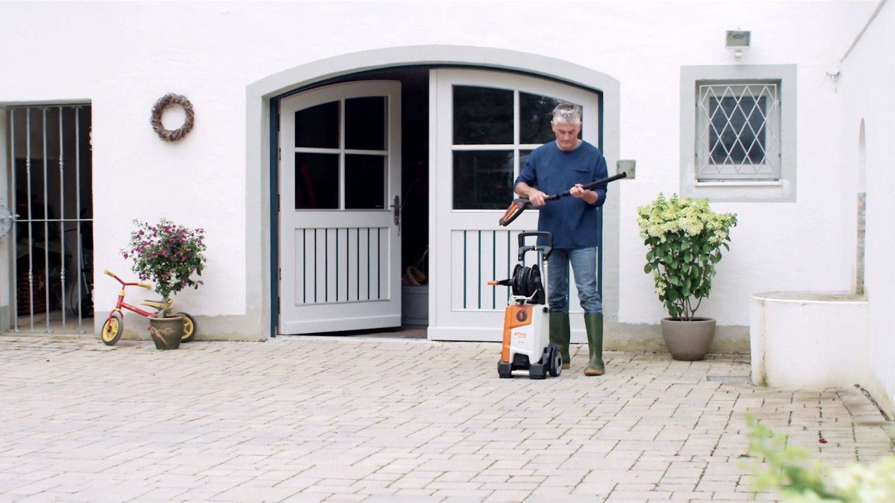 Čovek stoji ispred bele kuće i priprema STIHL uređaj za pranje pod visokim pritiskom za čišćenje fasade