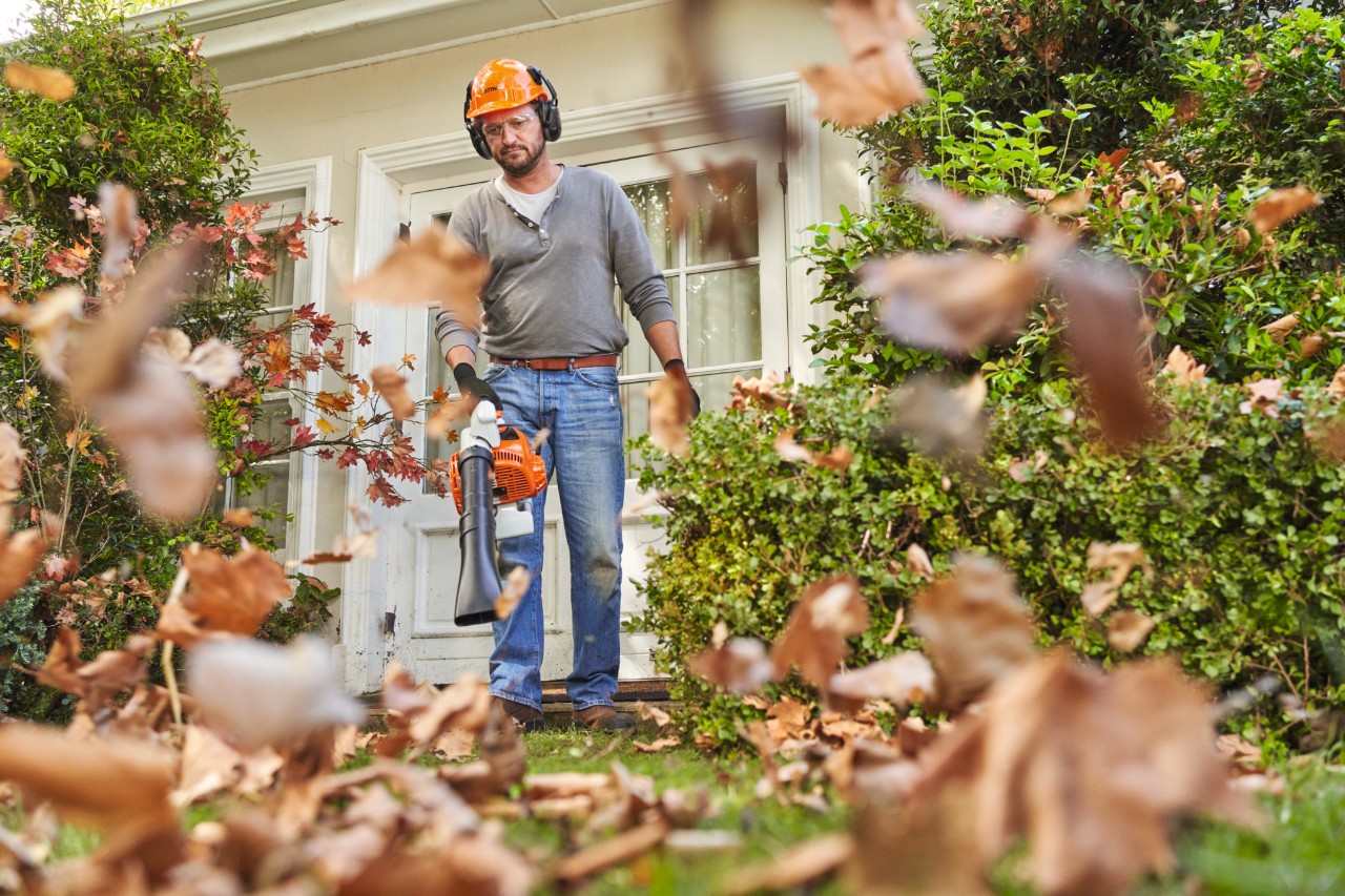 Čovek u STIHL zaštitnoj opremi ispred kuće oduvava lišće pomoću duvača