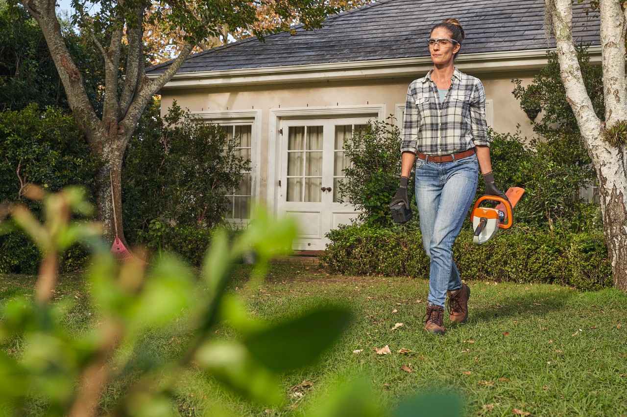 Žena sa STIHL makazama za živu ogradu šeta po travnjaku ispred kuće