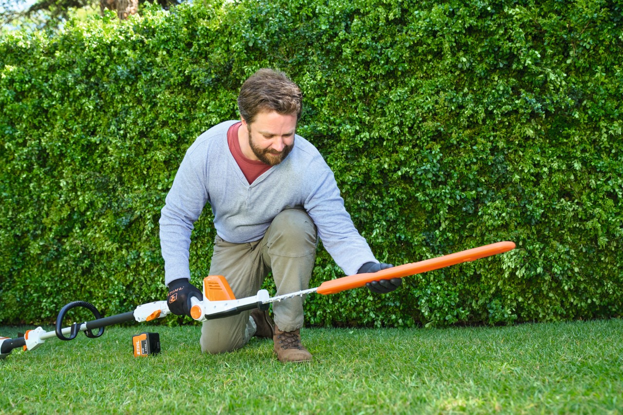 Čovek ispred žive ograde postavlja na travnjak baterijski trimer za živu ogradu STIHL HLA 56