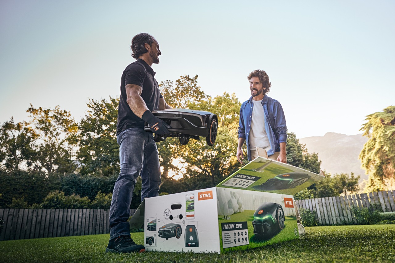STIHL stoji u vrtu klijenta kom želi da pomogne oko montaže i drži iMOW® EVO u rukama, dok je ispred njega kutija za iMOW®