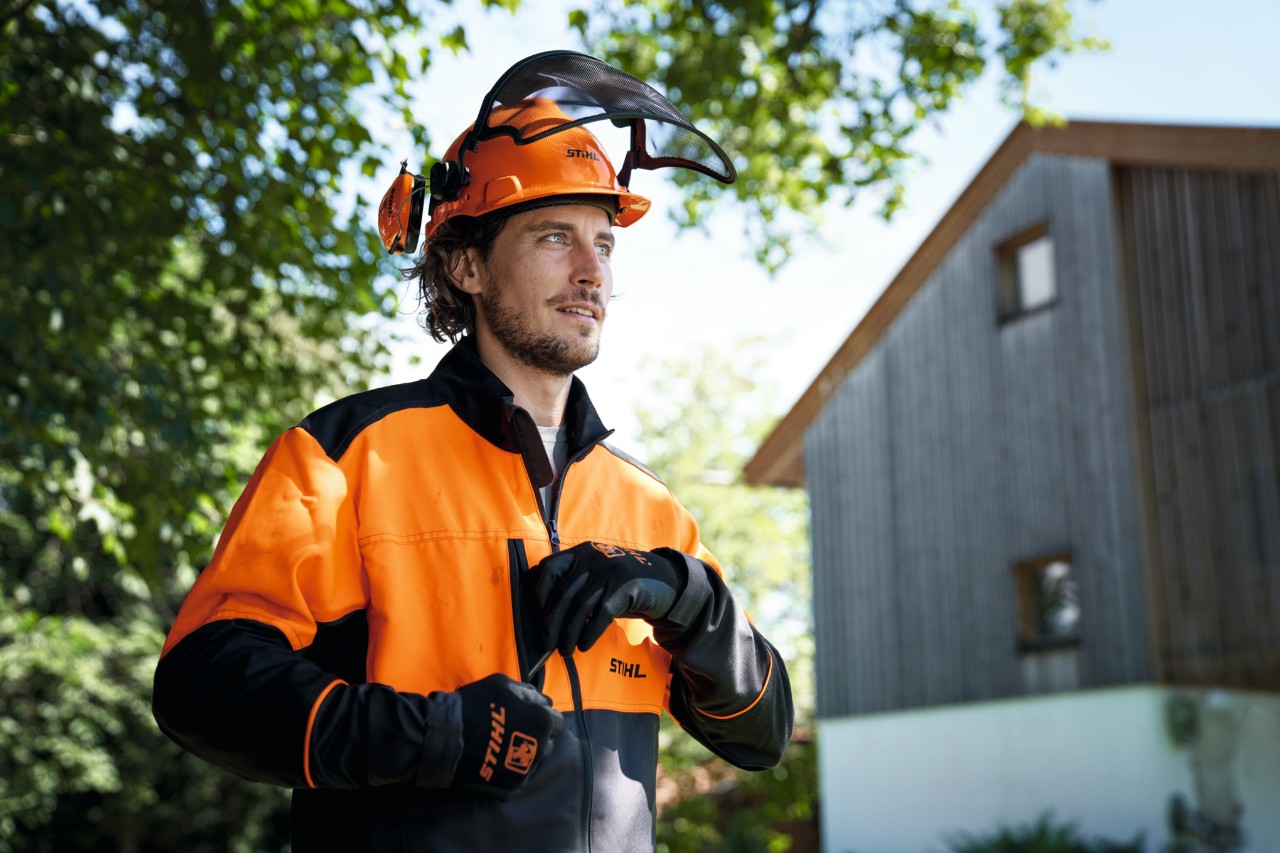 Samouveren čovek u narandžasto-crnoj STIHL zaštitnoj opremi sa kacigom i rukavicama, u pozadini se vide kuća i drveće 
